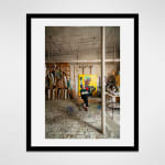 Framed photograph in black wood of an artist, Carmen Cicero, sits in front of a large figural painting in an industrial loft space filled with stored paintings and materials