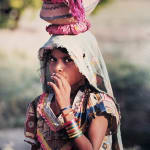 Pphotograph of a young woman with her hand raised to her mouth and fabric draped oved her head as she carries a vessel on her head