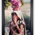 Framed photograph of a young woman with her hand raised to her mouth and fabric draped oved her head as she carries a vessel on her head
