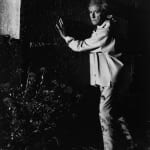 Black and white photograph of Jean Cocteau standing in grass with his hands against a wall of a building