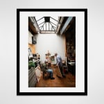 Framed photograph in black wood of artist Ken Jacobs standing and artist Flo Jacobs sitting at an easel with an unfinished painting in a loft setting filled with books and illuminated by a skylight