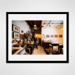 Black framed photograph of an artist, Joe Haske, leans against a worktable in a loft setting filled with materials and paintings