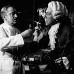 Black and white photograph of Henri Crémieux in a white coat lighting a cigarette for Jean Cocteau dressed as the poet in 18th-century nobleman costume