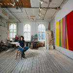 An artist, Betsy Kaufman, sits in an airy, sunlit loft setting surrounded by colorful abstract paintings