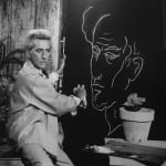 Black and white photograph of Jean Cocteau outside sitting and holding his self-portrait done on a black canvas with a white outline of his face