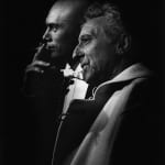 Black and white profile close-up photograph of Yul Brynner smoking a cigarette behind Jean Cocteau where both men are looking to the left
