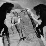 Black and white photograph of Jean Cocteau lying on a sheet outside lined with bricks between two horse-monster shadows which curve into Cocteau’s body