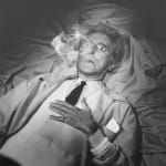 Black and white photograph of Jean Cocteau with glass eyes lying down on a wrinkled bed sheet with smoke coming out of his mouth