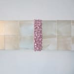 Detail of a row of white iridescent cubes made of acrylic paint on MDF with a pale pink textured strip at its center is mounted on a white wall.