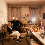 two married arists, Nels and Carol Pierce sitting in a studio in a loft setting surrounded by drawings, musical instruments, and materials