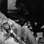 Black and white photograph of Jean Cocteau sleeping on the ground as a black horse looks down at him