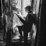 Black and white photograph of Andy Warhol reading the newspaper next to an open door in a foil-lined room