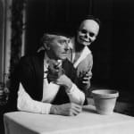 Black and white photograph of Jean Cocteau holding a flower and sitting at an empty table with an empty flower pot with Edouard Dermith with skeleton makeup standing behind him