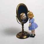 The image shows a charming vignette featuring a small figurine of a young girl in a blue dress looking at her reflection in an oval-shaped antique brass mirror. The doll has blond hair, a blue dress, and black shoes, and she appears to be engaged with her