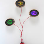The image displays a contemporary artwork featuring three circular elements mounted on the wall, each containing a large, realistic eye with different colored irises—one green, one yellow, and one purple. The eyes are connected by red wires that originate