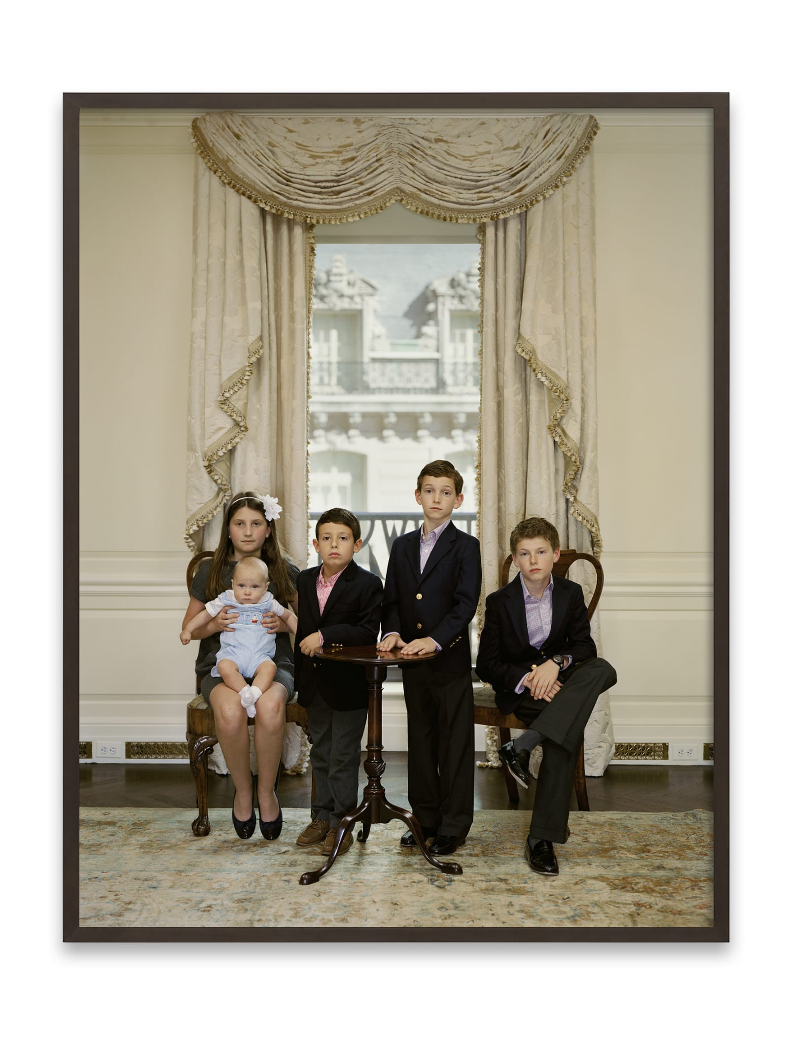 A Rineke Dijkstra portrait of 5 children: 3 boys and a girl holding a baby, in front of a window with an elegant beige curtain.