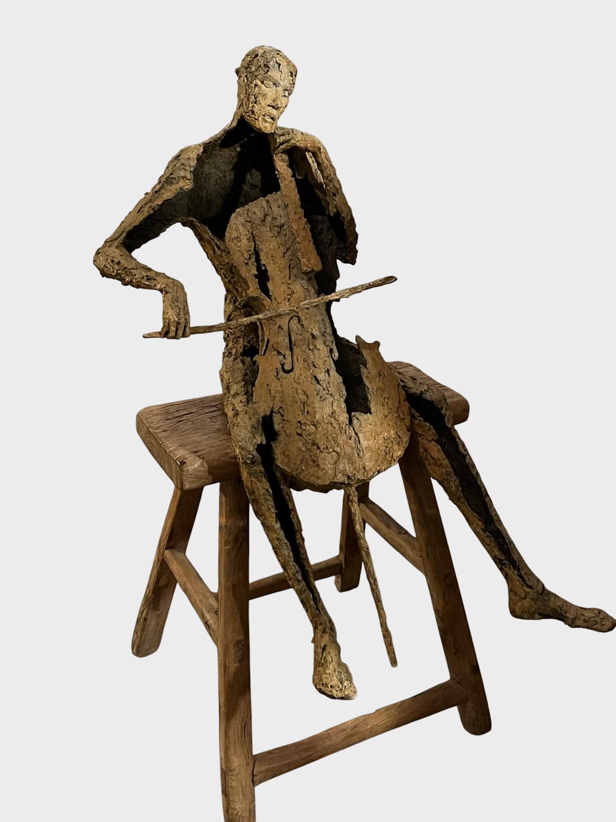 Bronze sculpture ‘Le Violoncelliste Assis’ by Jacques Van den Abeele, depicting an elongated seated musician playing the cello, with carved, textured surfaces typical of the artist.