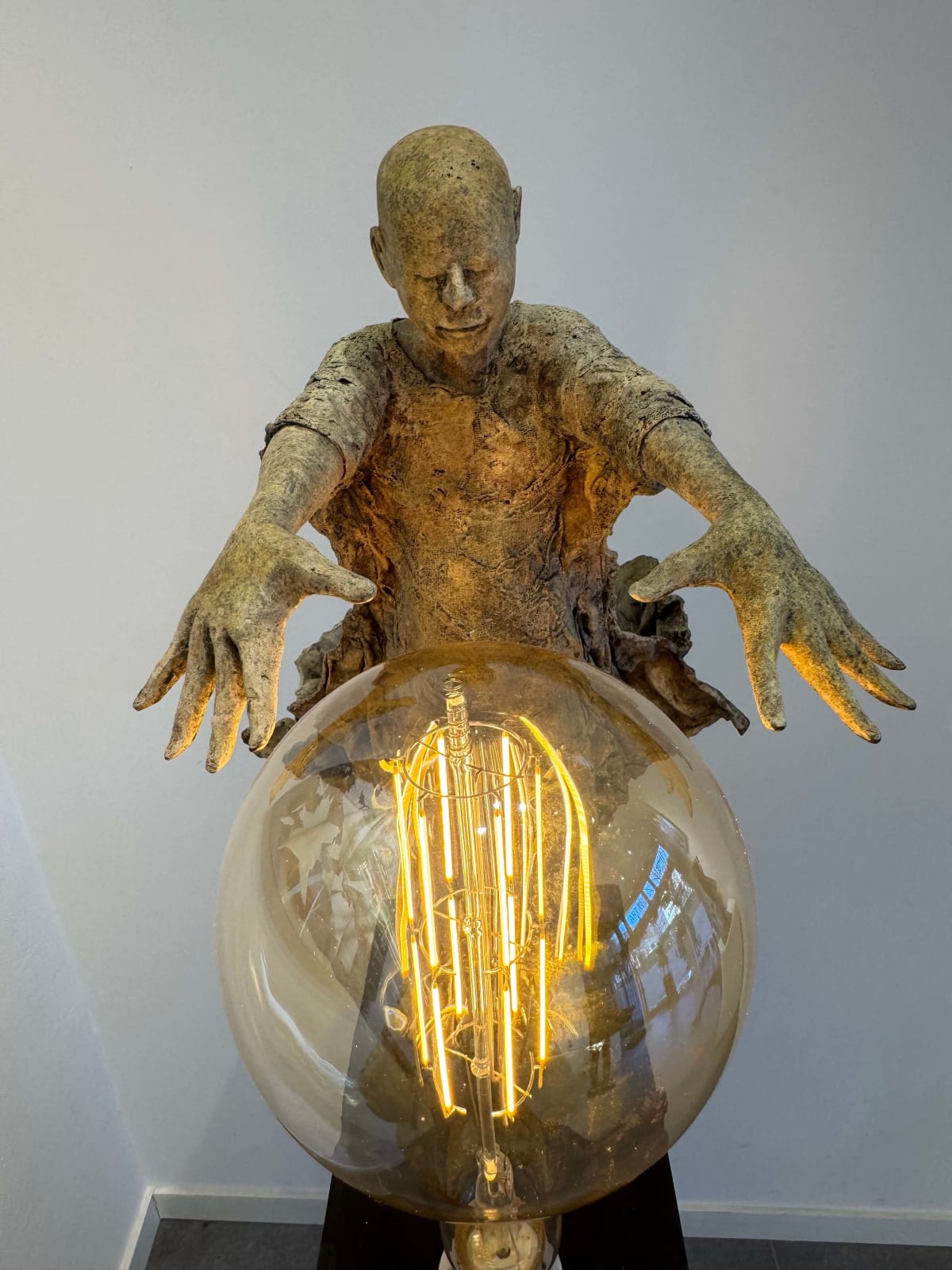Visionary XL met lichtgevende jongen sculptuur, bronzen sculptuur van Lieven d'Haese, tuinsculptuur van een jongen die zijn armen opent om een ​​gloeilamp in zijn handen aan te steken met verbeeldingskracht, magie, droom, hedendaagse kunstsculptuur, licht, interieurontwerp in de Art Yi galerie in Brussel.