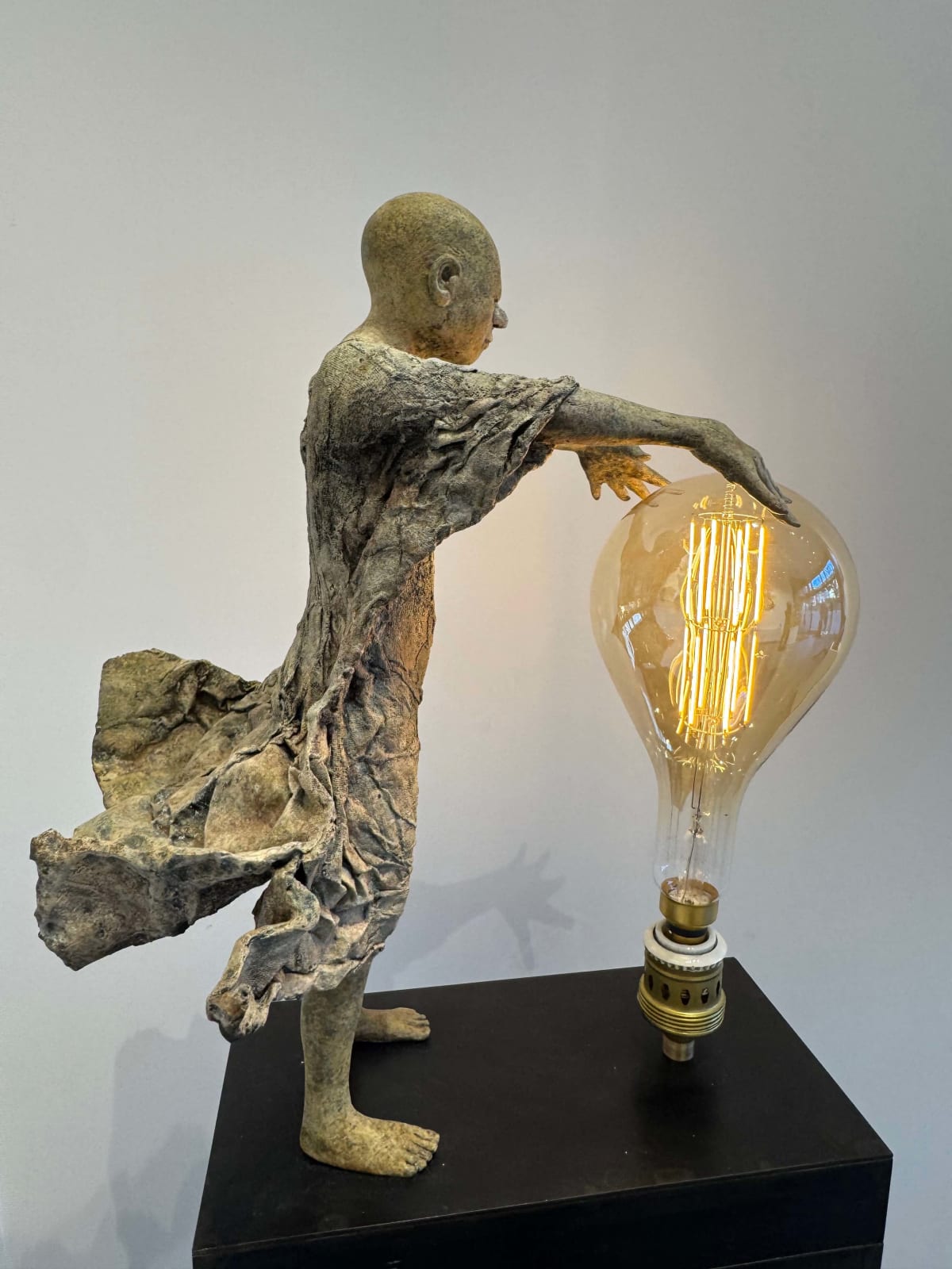 Visionary XL met lichtgevende jongen sculptuur, bronzen sculptuur van Lieven d'Haese, tuinsculptuur van een jongen die zijn armen opent om een ​​gloeilamp in zijn handen aan te steken met verbeeldingskracht, magie, droom, hedendaagse kunstsculptuur, licht, interieurontwerp in de Art Yi galerie in Brussel.