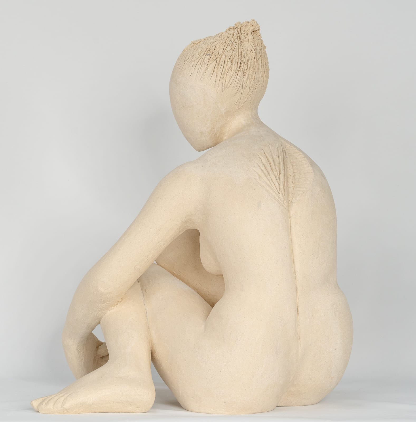 Seated white ceramic figure is looking over her left shoulder with arms draped around her knees which are tucked together.