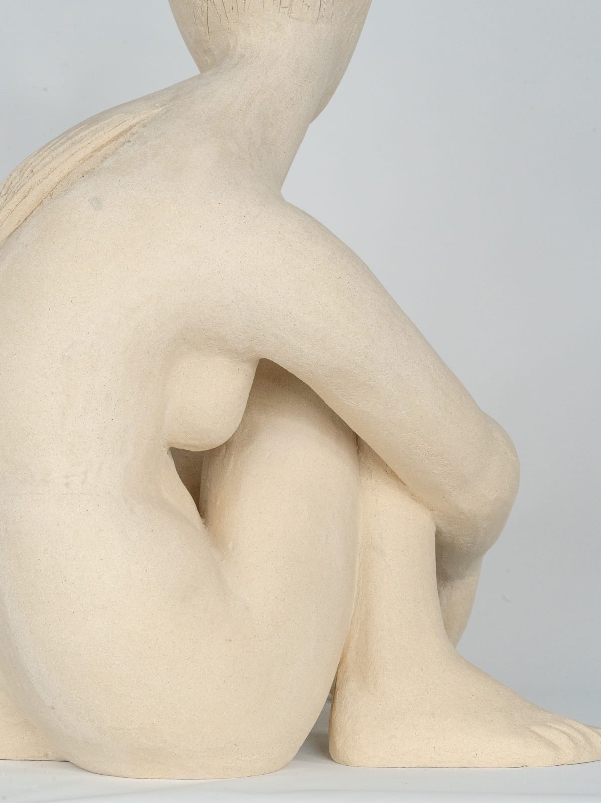 Seated white ceramic figure is looking over her left shoulder with arms draped around her knees which are tucked together.