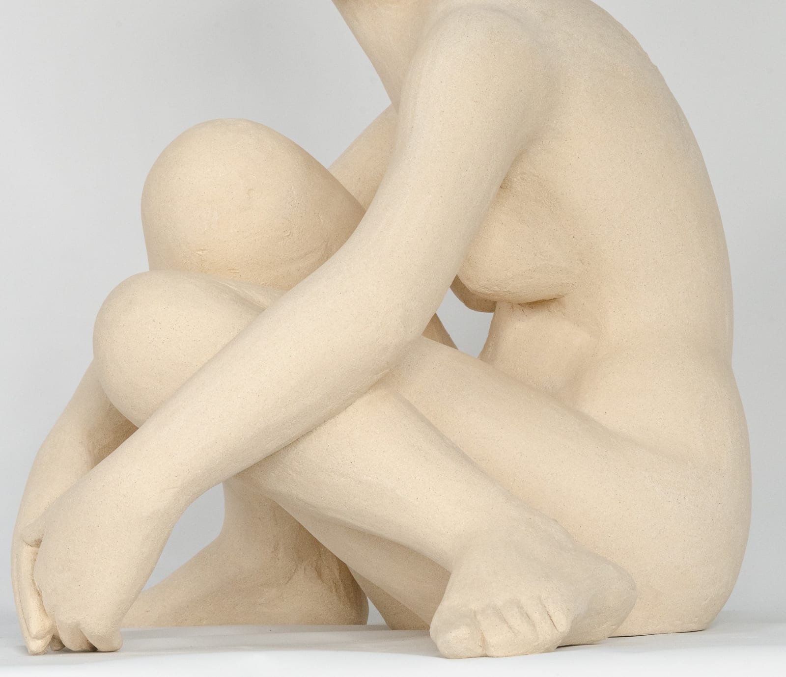 Seated white ceramic figure is looking over her left shoulder with arms draped around her knees which are tucked together.