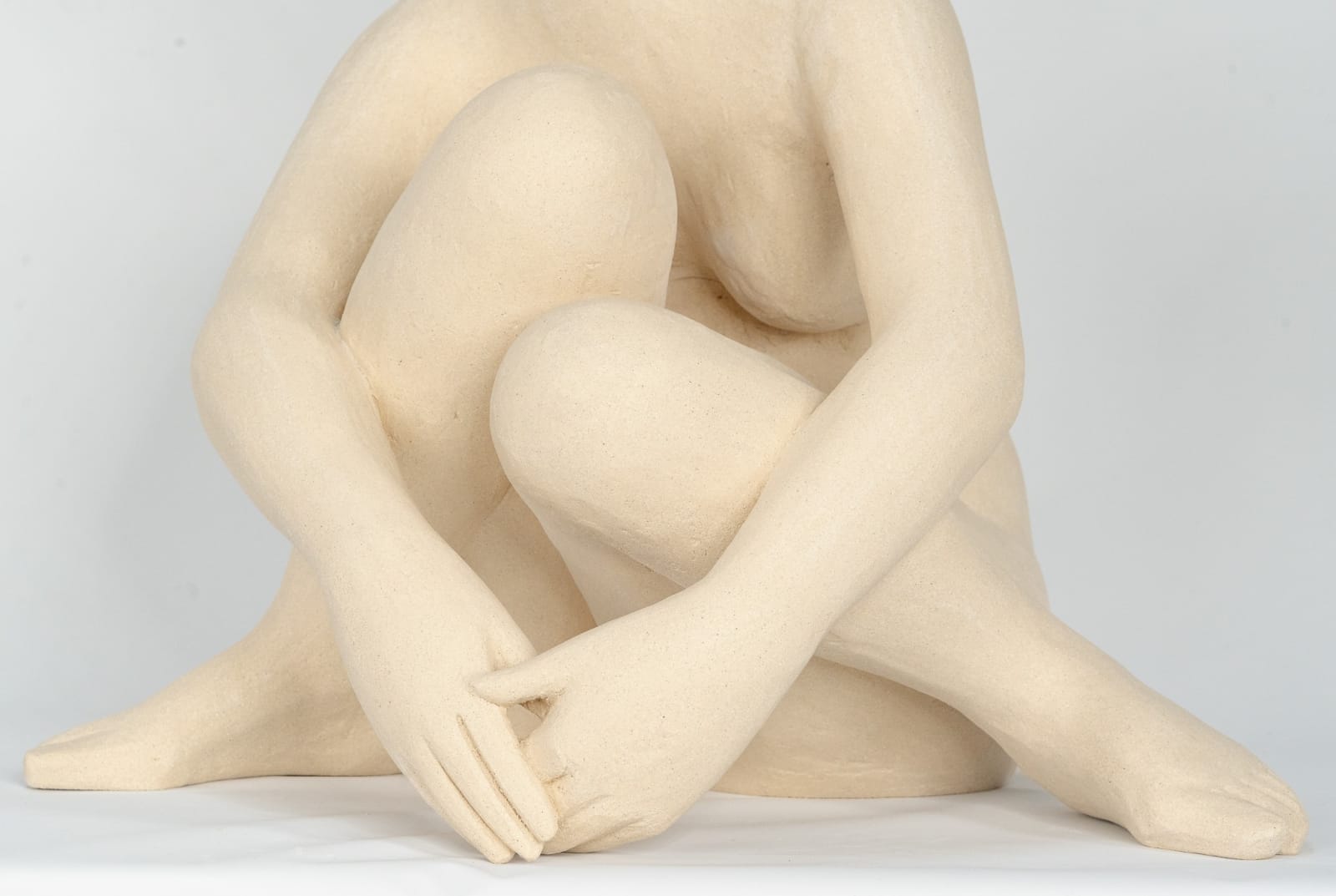 Seated white ceramic figure is looking over her left shoulder with arms draped around her knees which are tucked together.