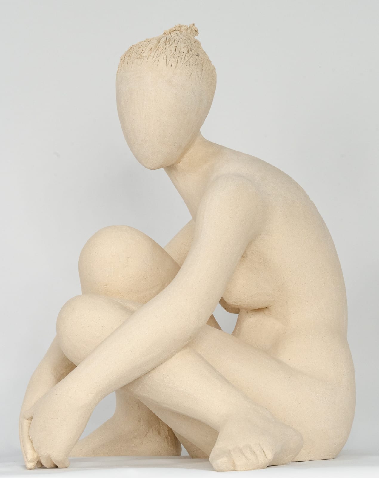 Seated white ceramic figure is looking over her left shoulder with arms draped around her knees which are tucked together.