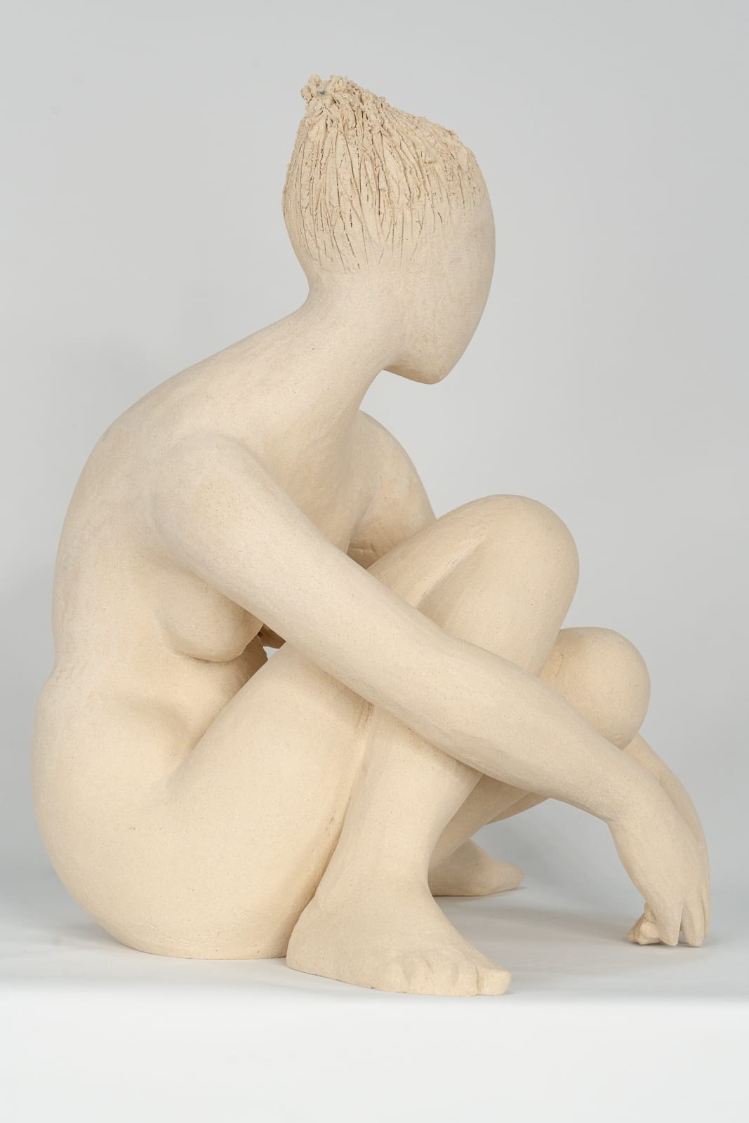 Seated white ceramic figure is looking over her left shoulder with arms draped around her knees which are tucked together.