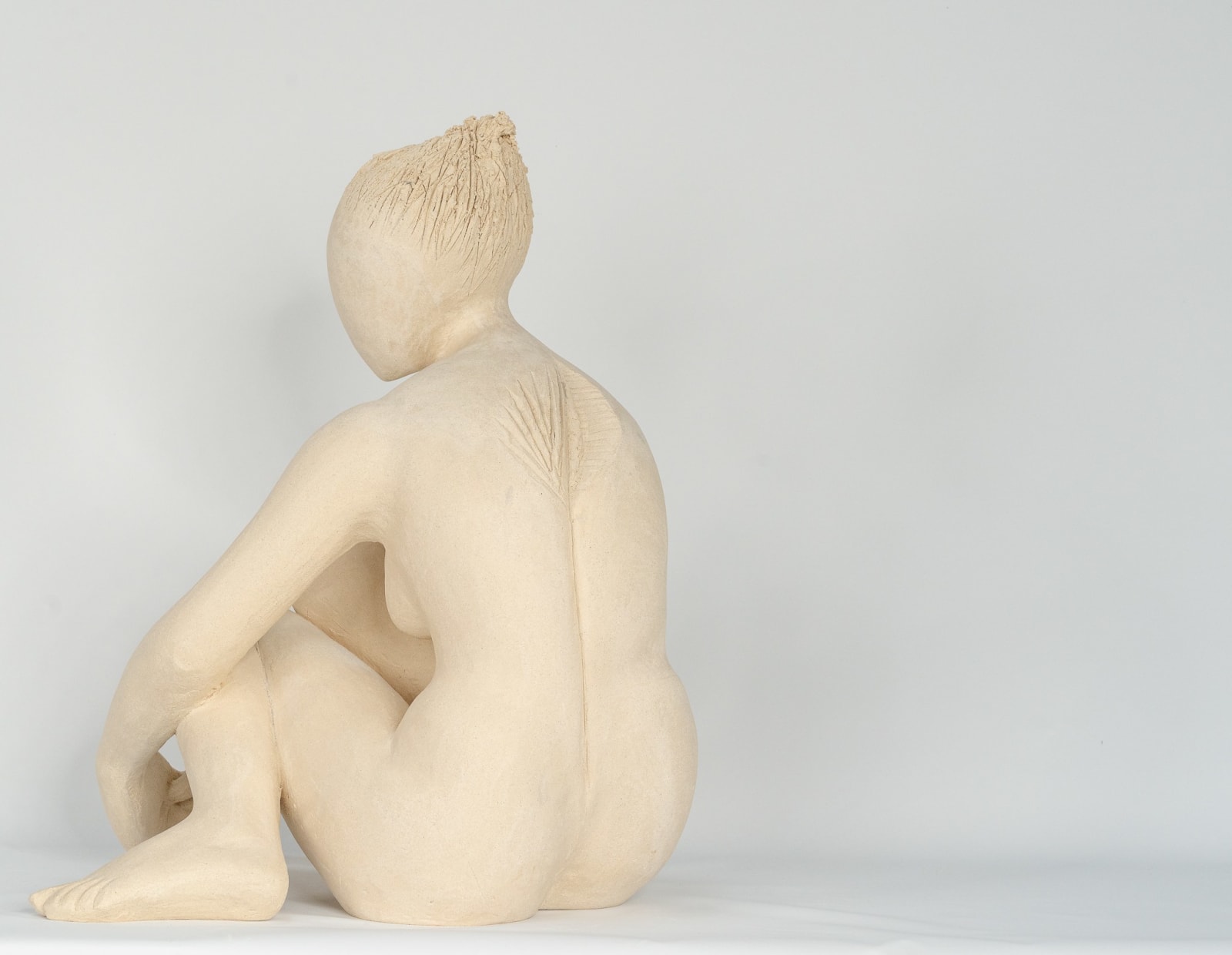 Seated white ceramic figure is looking over her left shoulder with arms draped around her knees which are tucked together.