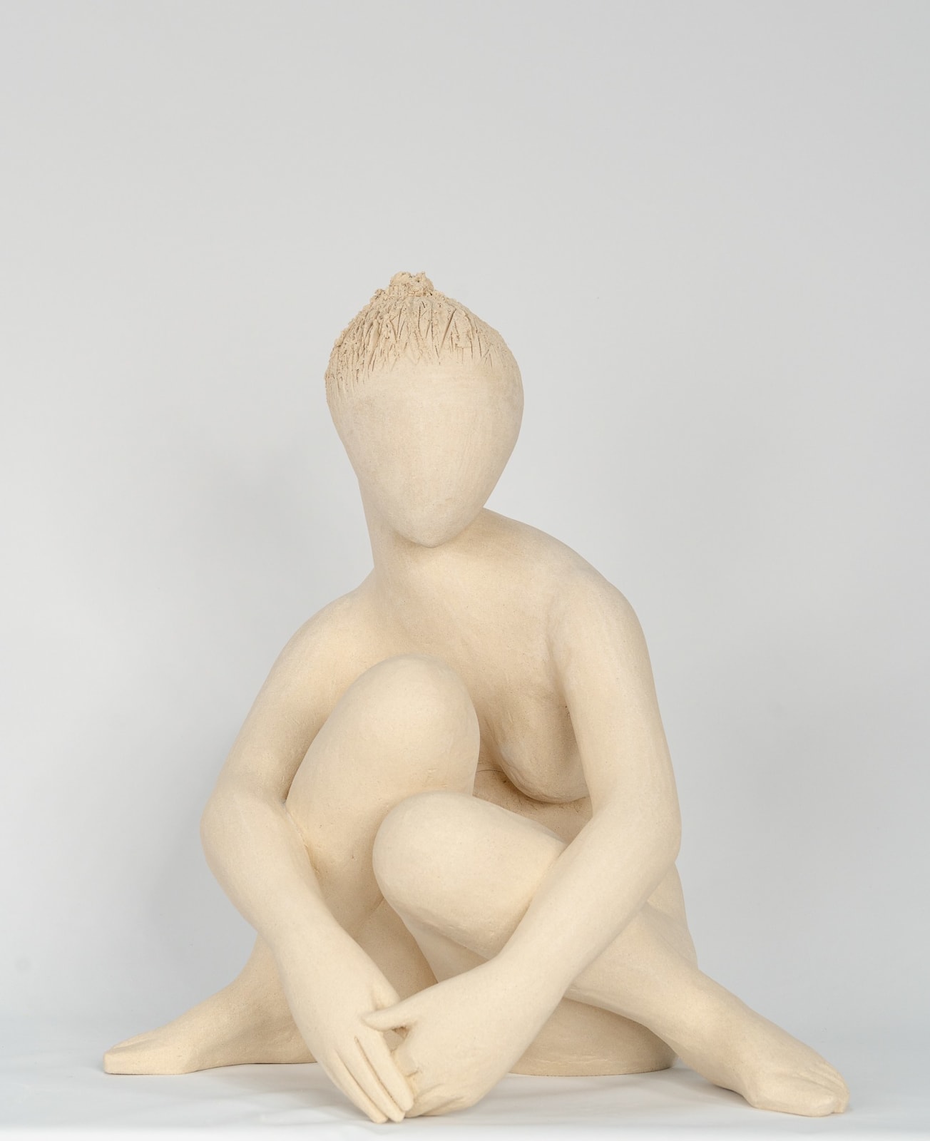 Seated white ceramic figure is looking over her left shoulder with arms draped around her knees which are tucked together.