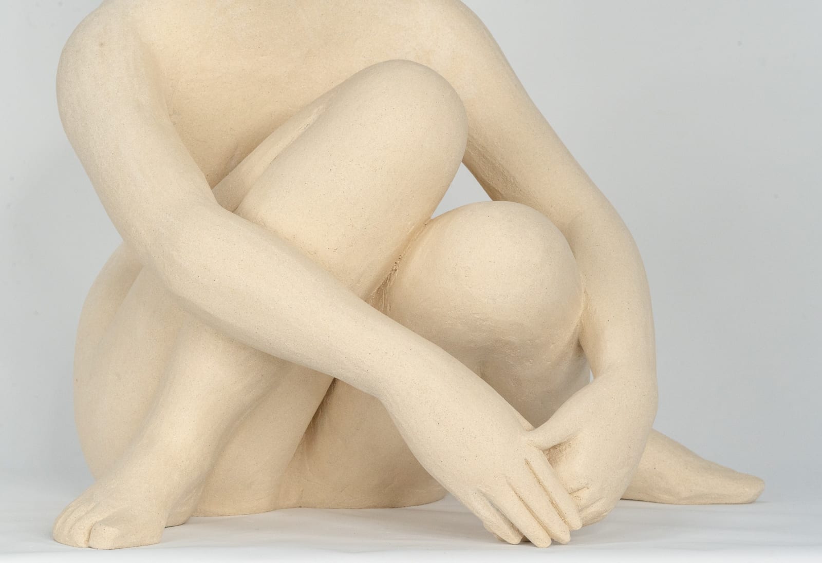 Seated white ceramic figure is looking over her left shoulder with arms draped around her knees which are tucked together.