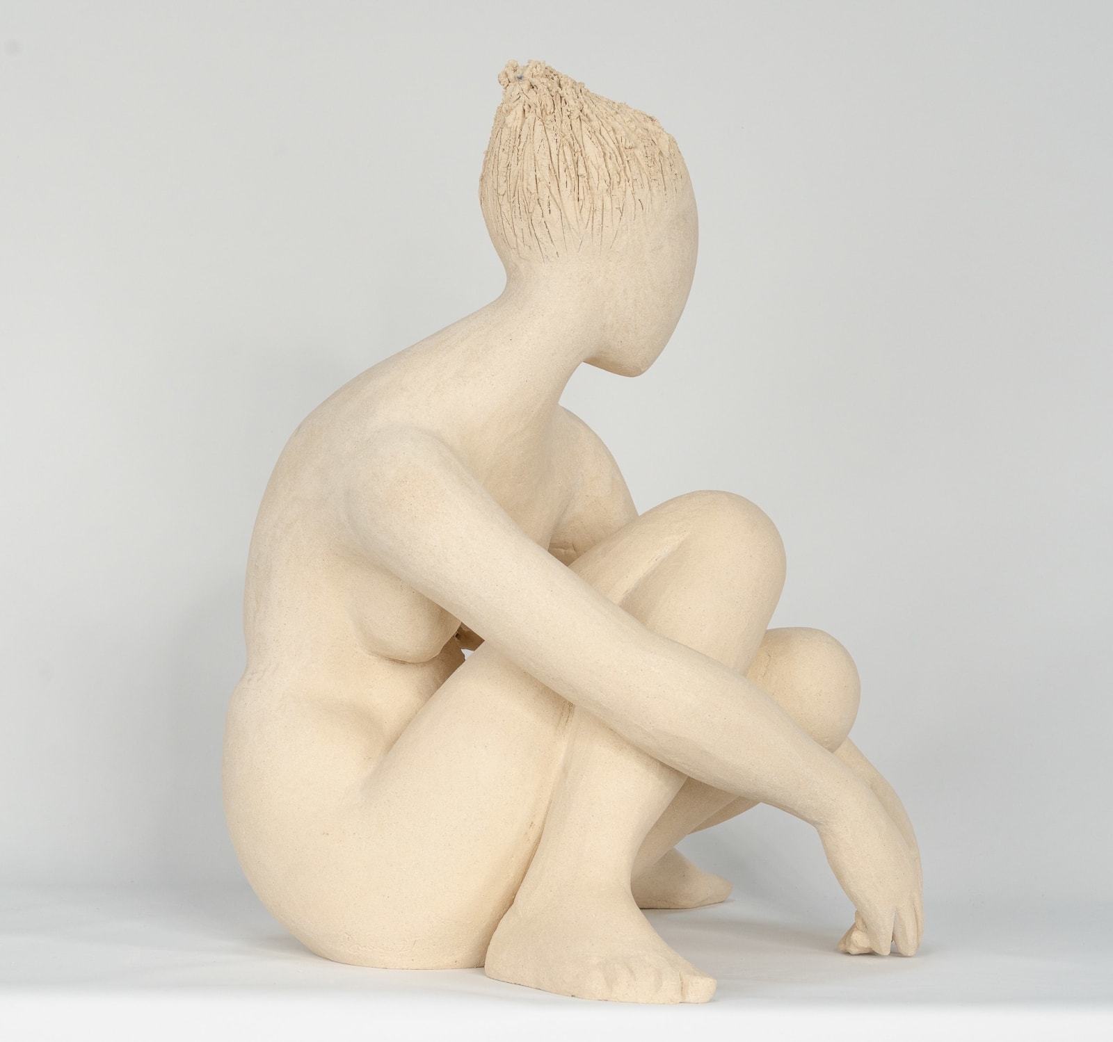 Seated white ceramic figure is looking over her left shoulder with arms draped around her knees which are tucked together.