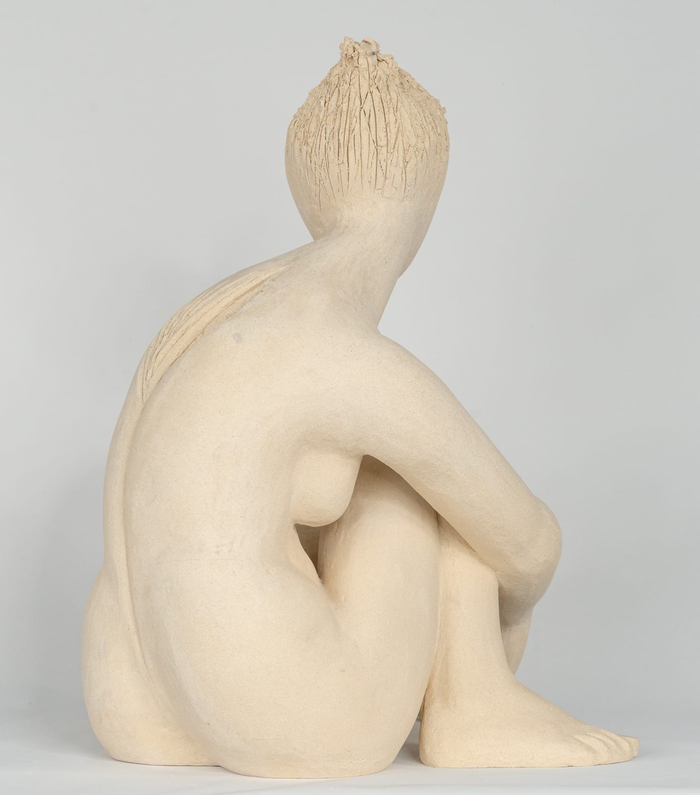 Seated white ceramic figure is looking over her left shoulder with arms draped around her knees which are tucked together.