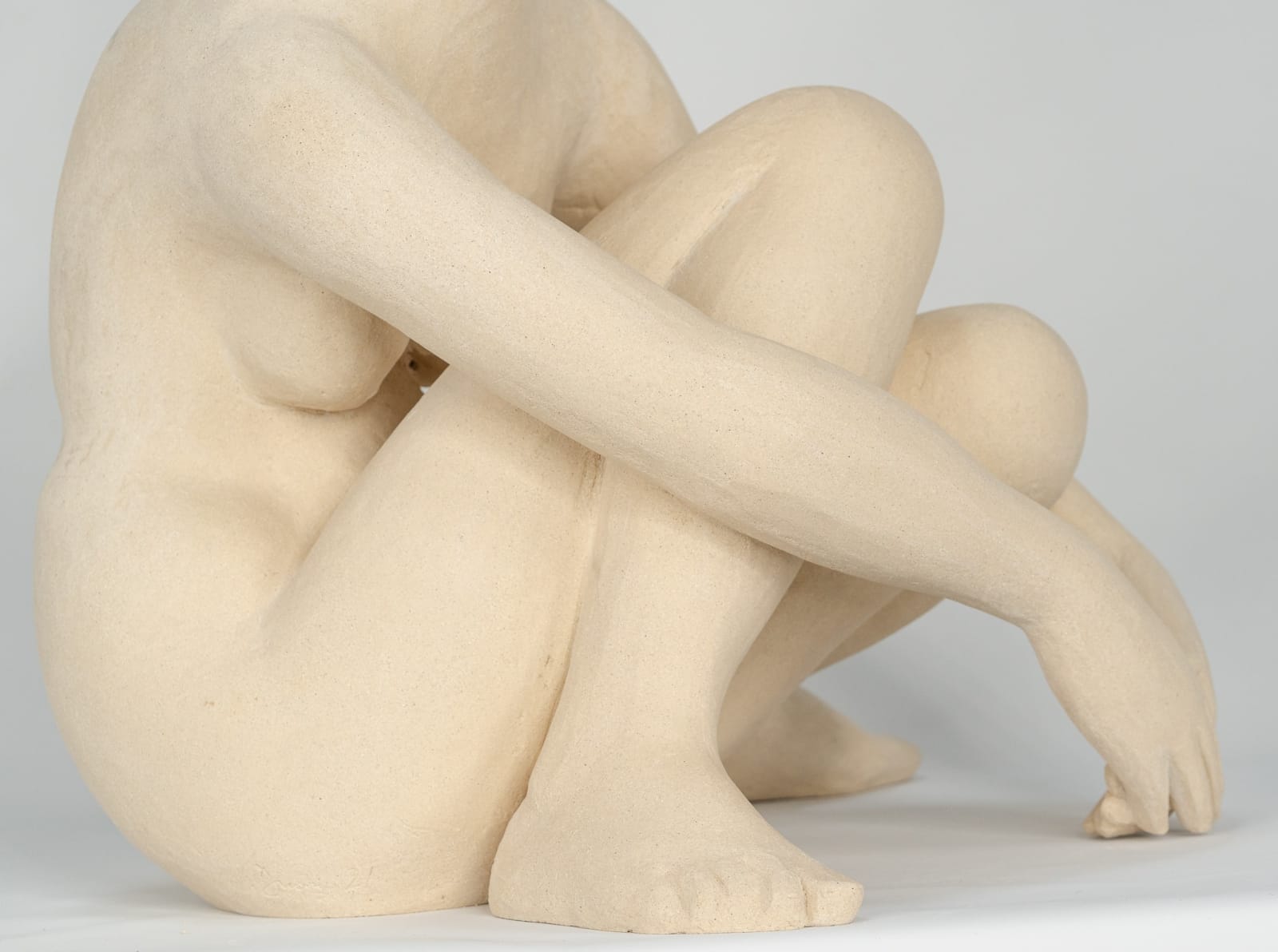 Seated white ceramic figure is looking over her left shoulder with arms draped around her knees which are tucked together.