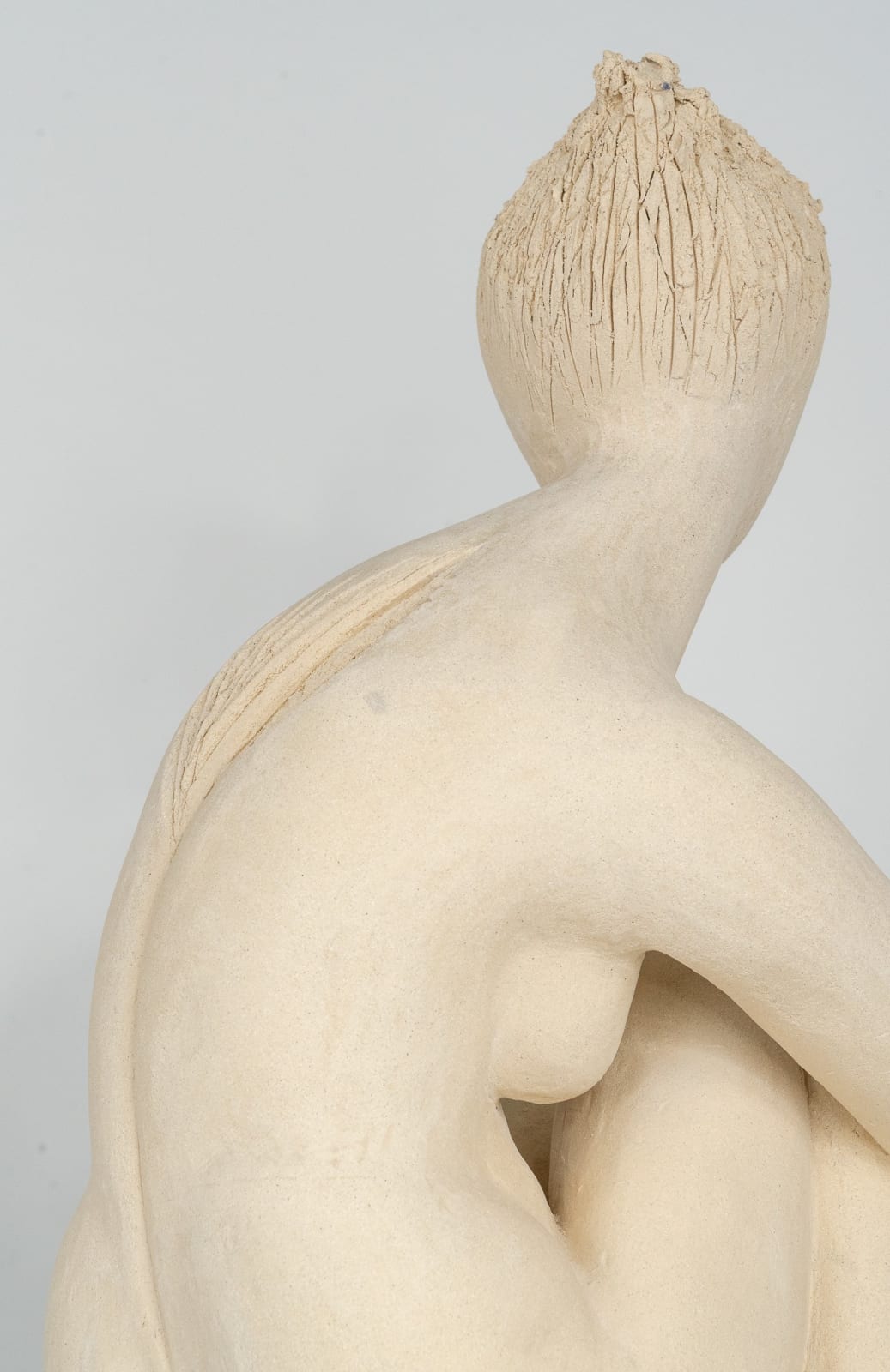Seated white ceramic figure is looking over her left shoulder with arms draped around her knees which are tucked together.