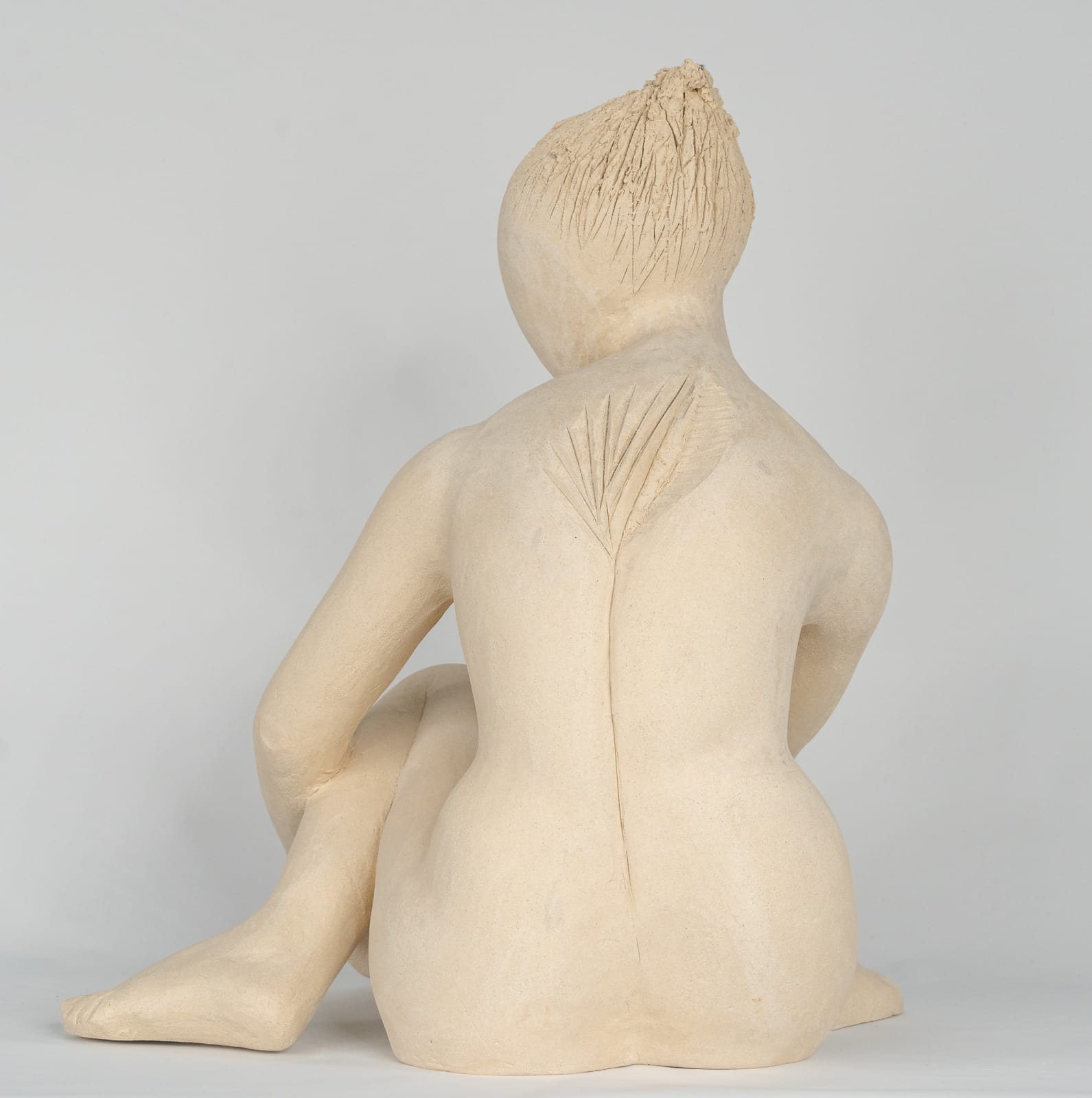 Seated white ceramic figure is looking over her left shoulder with arms draped around her knees which are tucked together.