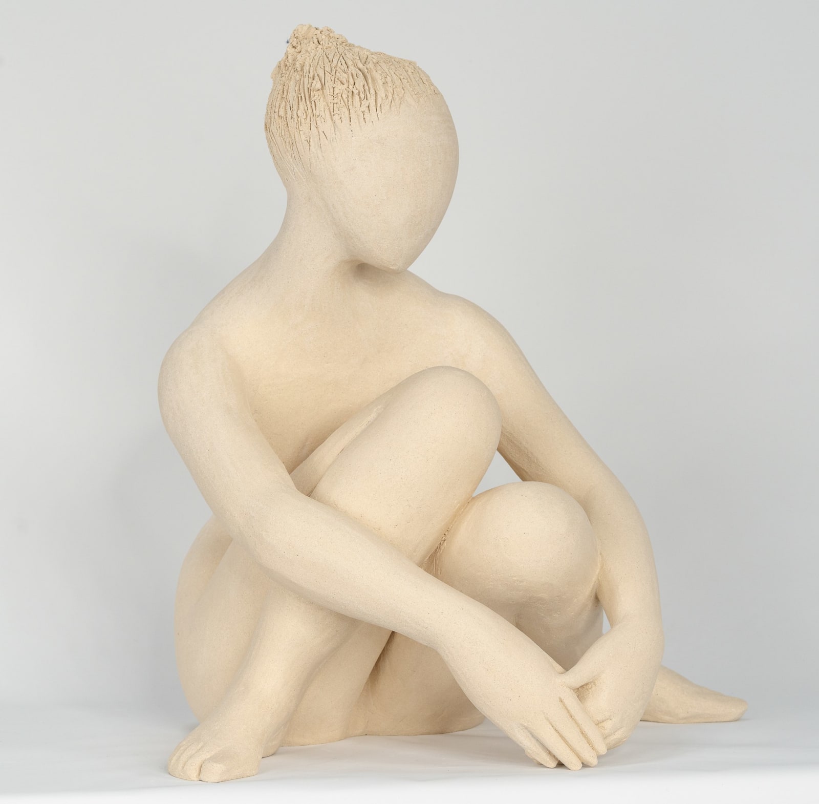 Seated white ceramic figure is looking over her left shoulder with arms draped around her knees which are tucked together.