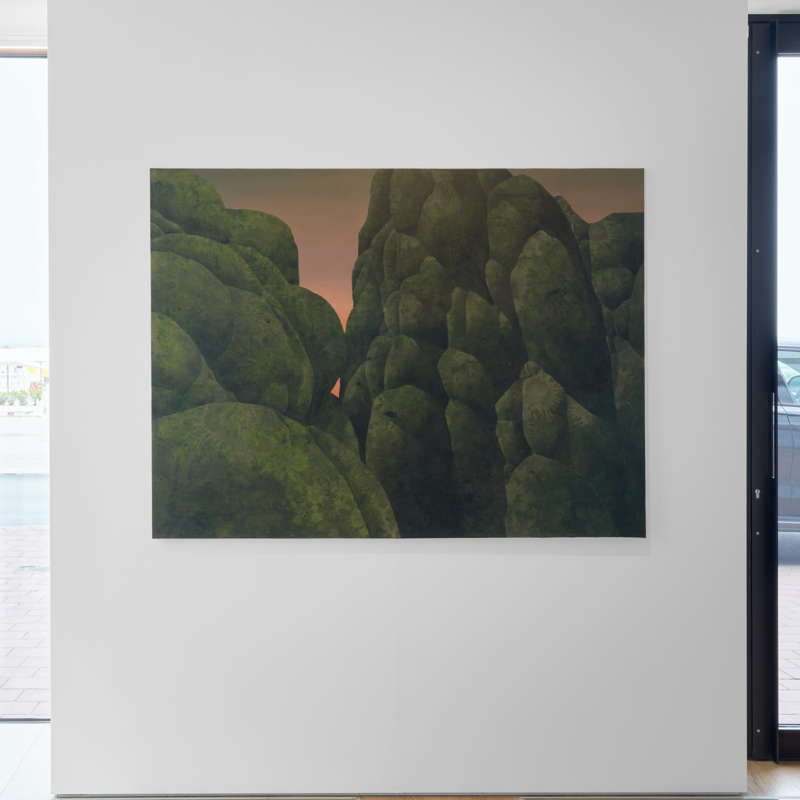 Painting titled “First Flora” (2023) by Silvia Giordani. The artwork features a dense arrangement of mossy green, organic forms resembling a surreal forest or mountain terrain, set against a soft gradient sky transitioning from warm pink to deep gray. Acrylic and oil on canvas. © Buysse Gallery.