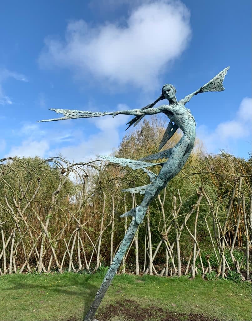A monumental bronze sculpture of a winged female stands in front of a willow walk at a Dorset sculpture park, Sculpture by the Lakes