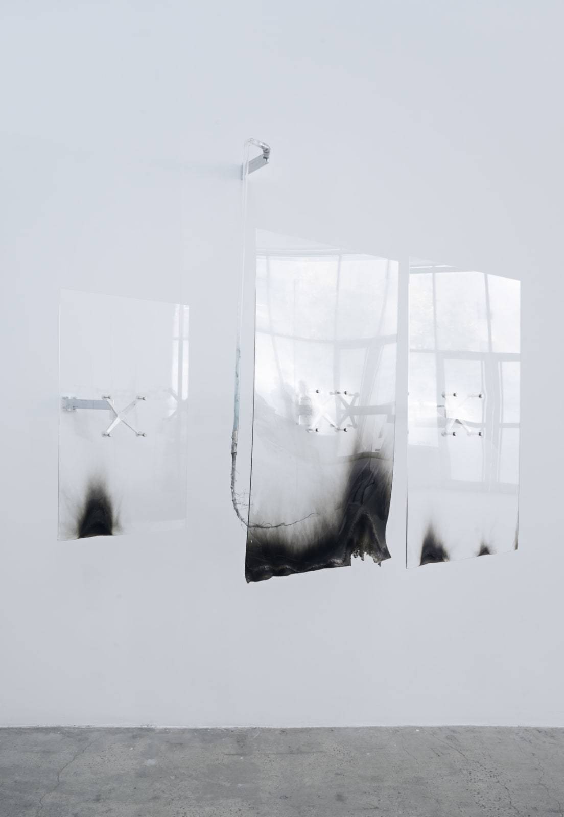 An image of three burned acrylic sheets attached to a white wall by aluminum spider brackets, suspended over a concrete floor.