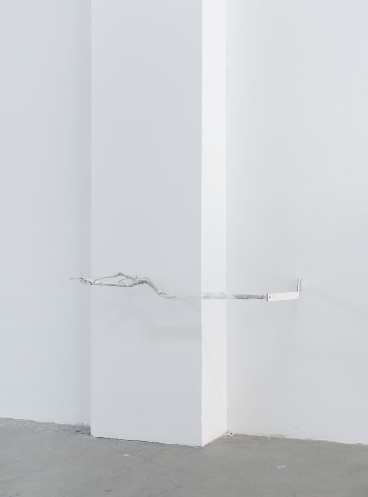An image of a pronged aluminum arm attached to a white wall by a white wall, suspended over a dark concrete floor.