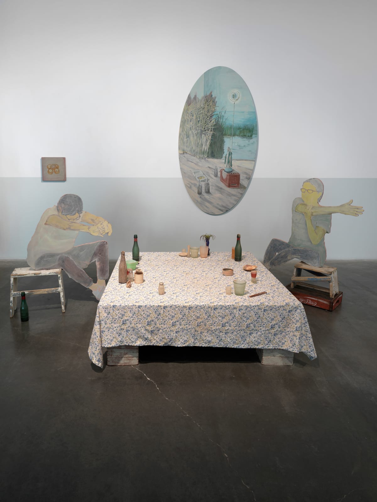 image of a mixed media installation consisting of two panels shaped and painted to depict people, posed at either end of a table covered in a floral cloth and bearing several small glasses and containers