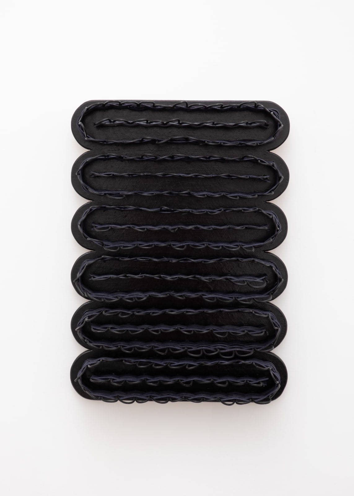 Wall-mounted sculpture consisting of a black wooden rectangular base with scalloped edges, with woven black reeds attached in a meandering pattern from end to end.