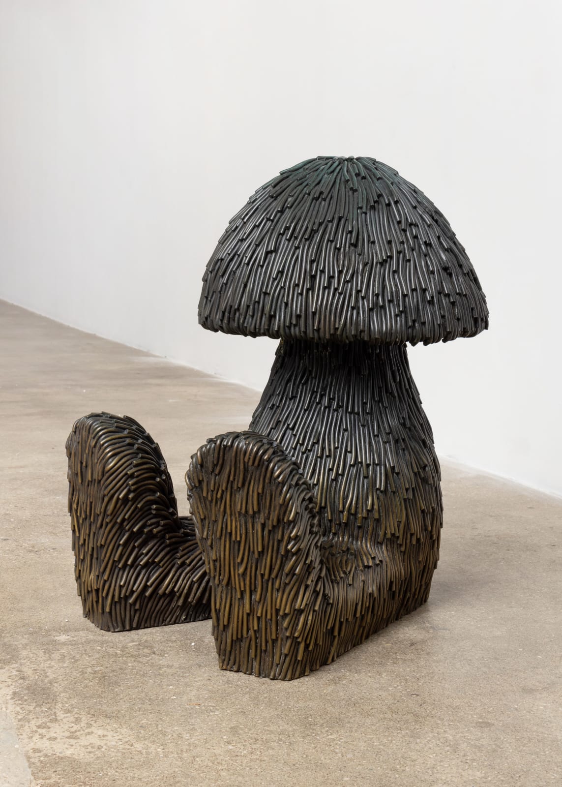 Free-standing bronze sculpture of a body with a mushroom-shaped head. The sculpture is cast with a surface texture resembling shaggy hair and treated with a greenish black patina that turns into bronze, from top to bottom. 