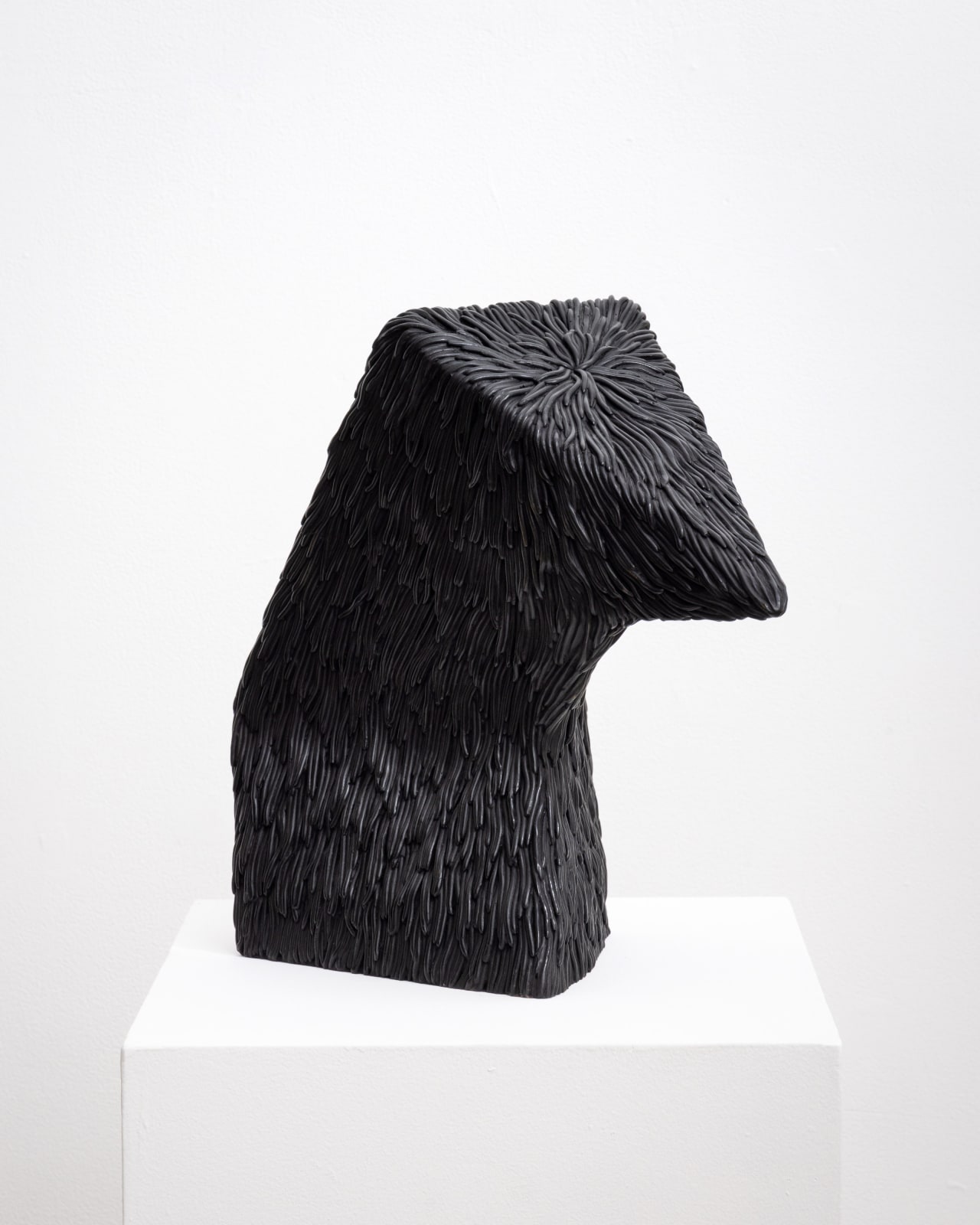 Free-standing bronze sculpture of an abstracted form with a flat triangular head that is bowing down. The sculpture is cast with a surface texture resembling shaggy hair and treated with a black patina. 