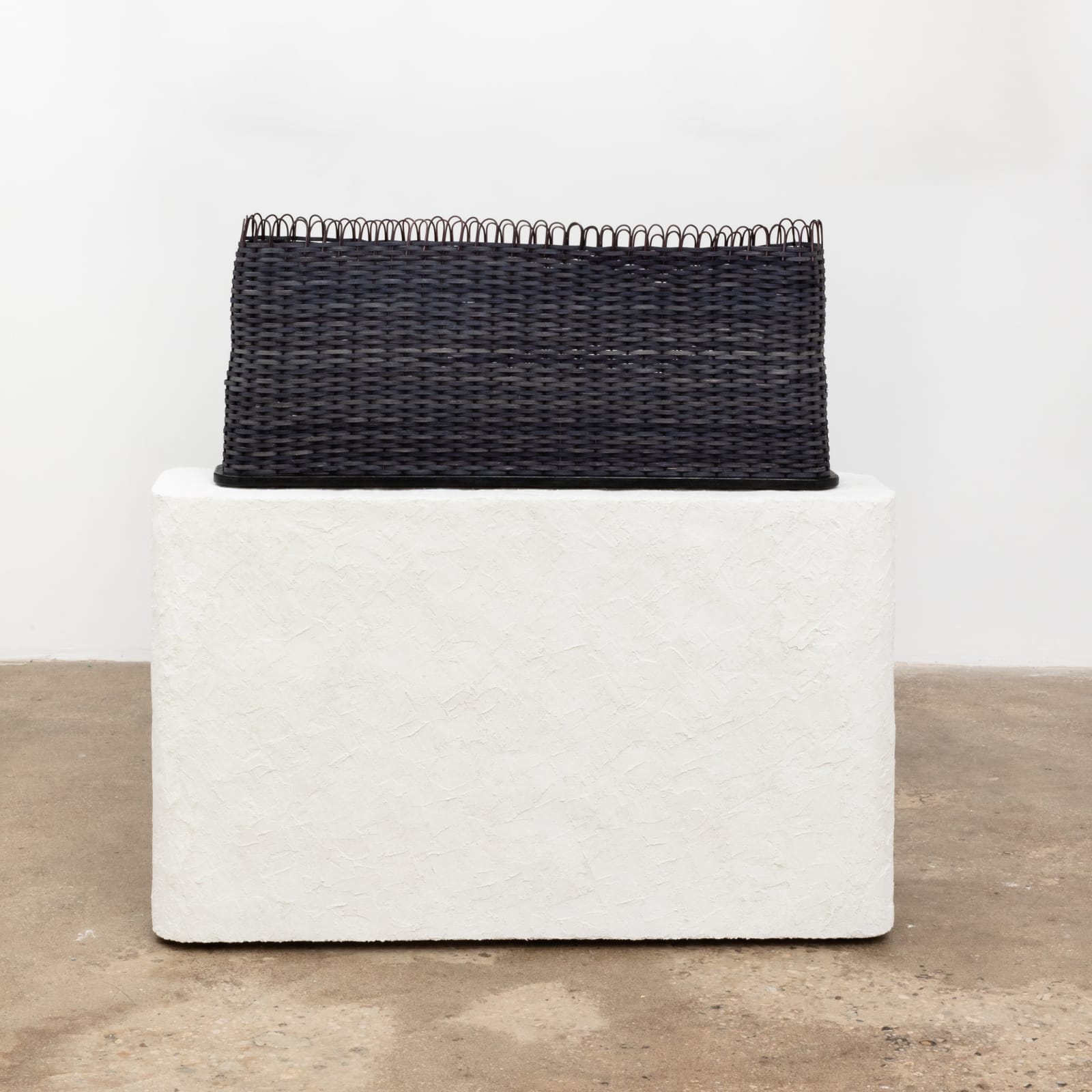 Free-standing sculpture woven with natural dyed, black reed to resemble an elongated basket, which sits atop a similar shaped white stucco pedestal base. 