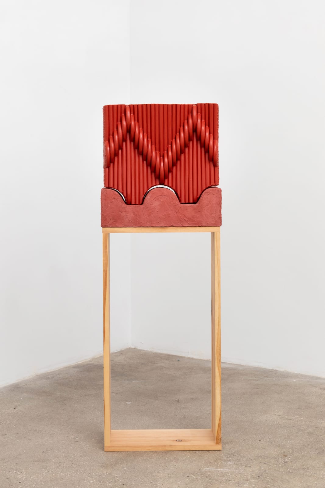 Free-standing sculpture consisting of carved polystyrene pieces aggregated together to form a red hued, interlocking patterned square form that sits on top of a rectangular wooden pedestal. 