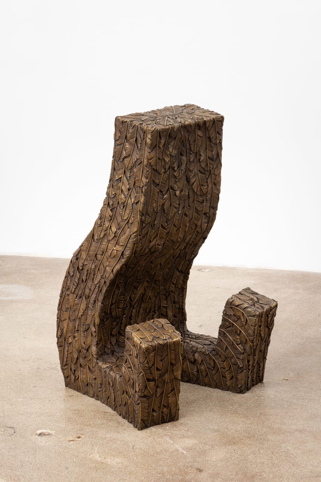 Free-standing bronze sculpture of a rectangular form sitting with outstretched legs. The sculpture is cast in surface texture resembling feathers and treated with a golden brown patina. 