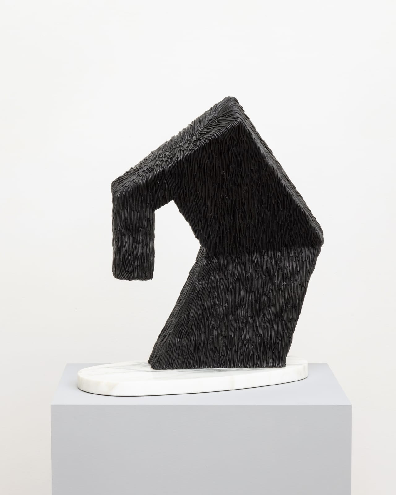 Free-standing bronze sculpture of a tall angular shape that is bending over and perched on a rounded marble base. The sculpture is cast with a surface texture resembling shaggy hair and treated with a black patina. 